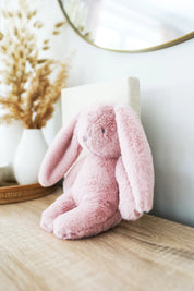 Alimrose Darcey Plush Baby Bunny 27cm in petal pink sitting on a wooden dresser in a nursery setting.