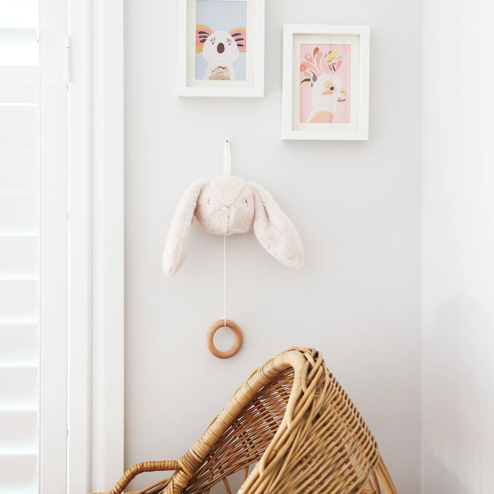 Alimrose Darcey Bunny Musical toy in ivory, hanging on nursery wall above wicker bassinet with framed artwork.