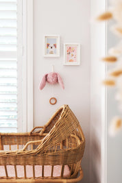 Alimrose Darcey Bunny Musical toy in petal pink, hanging on nursery wall above wicker bassinet with framed artwork.