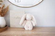 Alimrose Darcey Plush Baby Bunny 27cm in ivory displayed on a wooden dresser beside a baby keepsake box and vase.