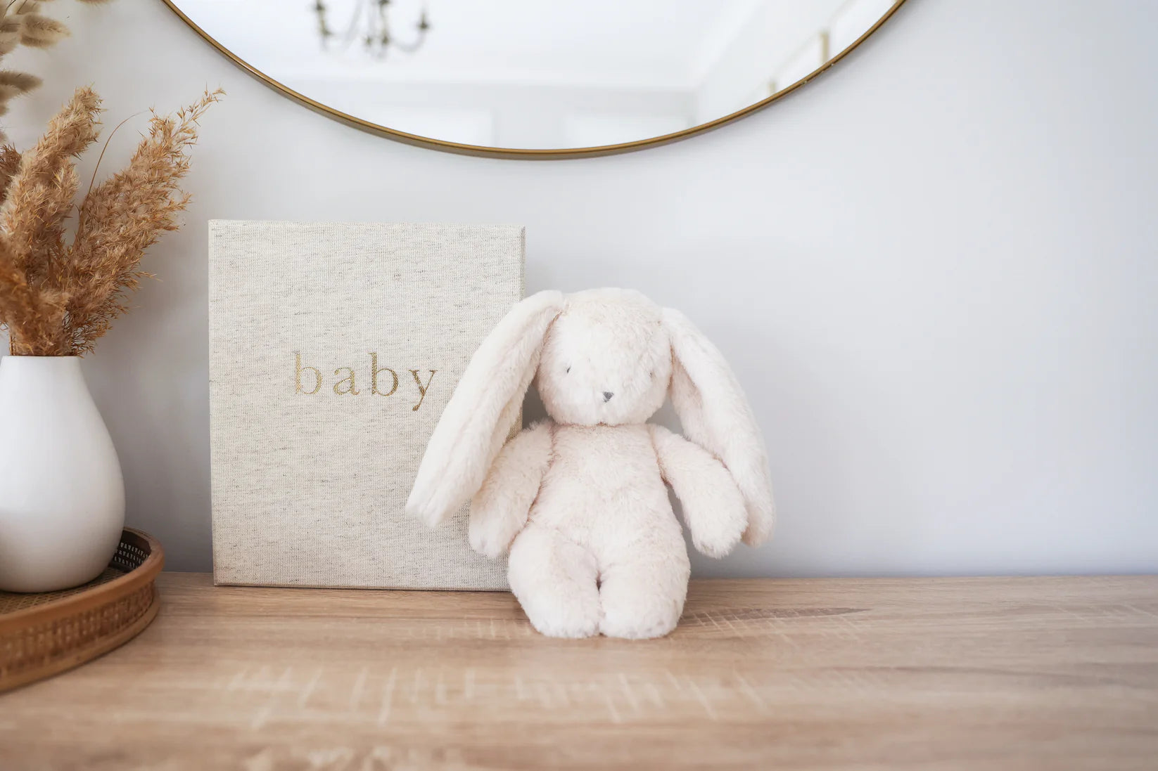 Alimrose Darcey Plush Baby Bunny 27cm in ivory displayed on a wooden dresser beside a baby keepsake box and vase.