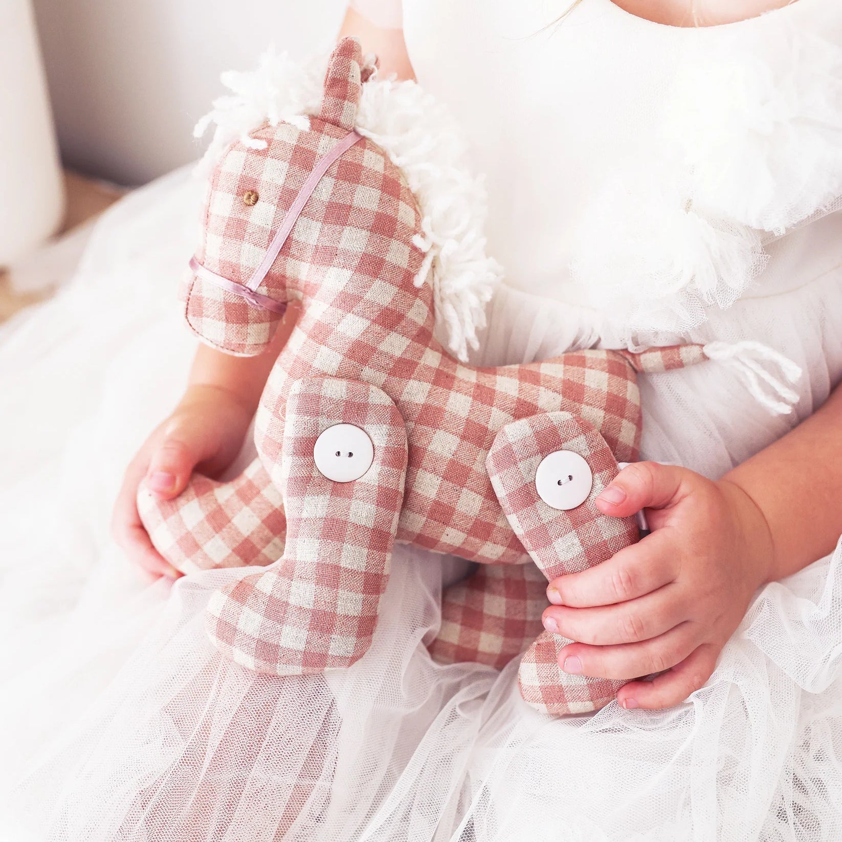 Child in white dress holding Alimrose Jointed Pony 22cm in rose check fabric with button joints.