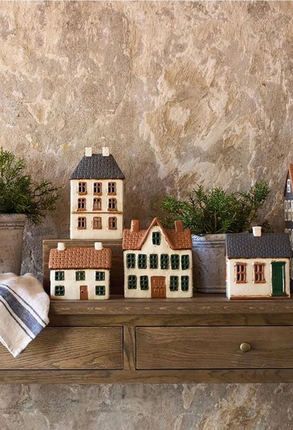 A closer view of ceramic tealight houses displayed on a rustic wooden table, including a tall townhouse, small cottages with red and brown roofs, and potted greenery in the background against a textured stone wall.