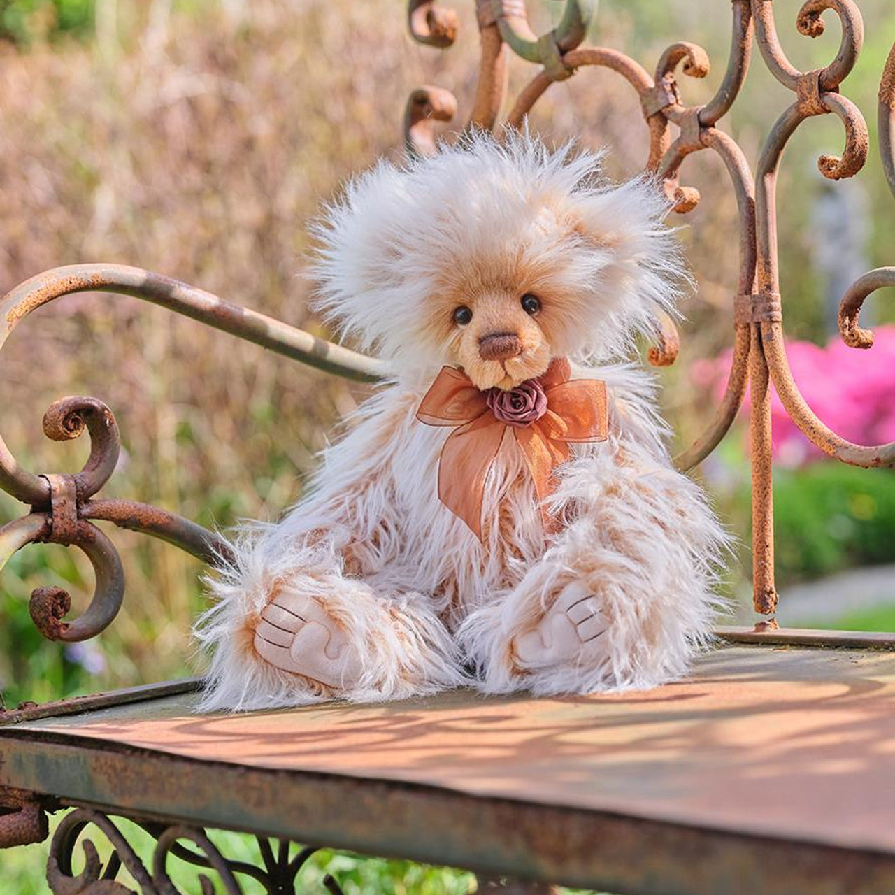 Charlie Bears Ari plush bear with cream and caramel fur and a brown organza bow, sitting on an ornate outdoor bench.