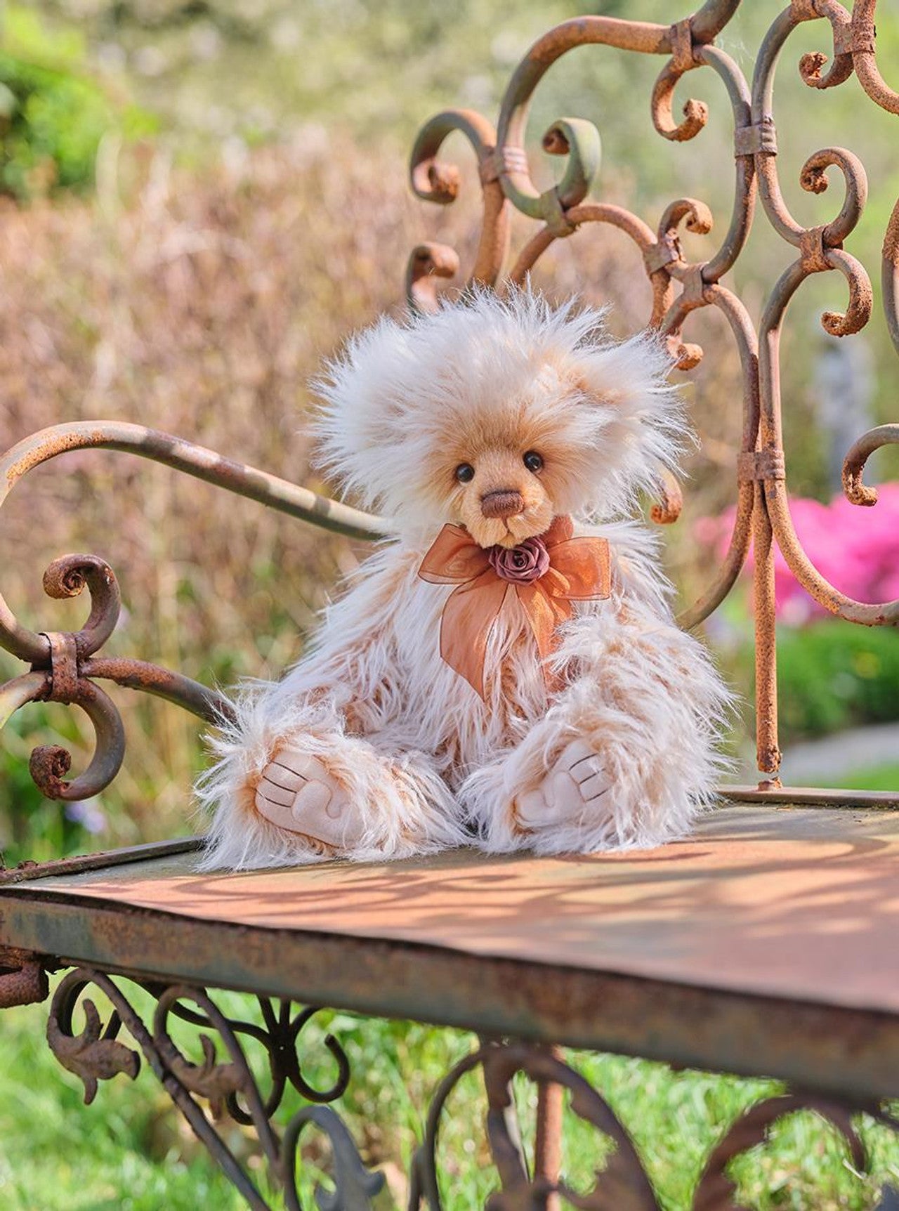 Charlie Bears Ari plush bear with cream and caramel fur and a brown organza bow, sitting on an ornate outdoor bench.