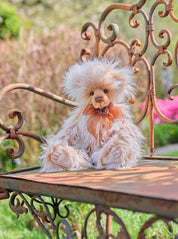 Charlie Bears Ari plush bear with cream and caramel fur and a brown organza bow, sitting on an ornate outdoor bench.