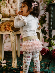 Toddler wearing white top and patterned leggings with gold waistband and pink frill tutu detail at back