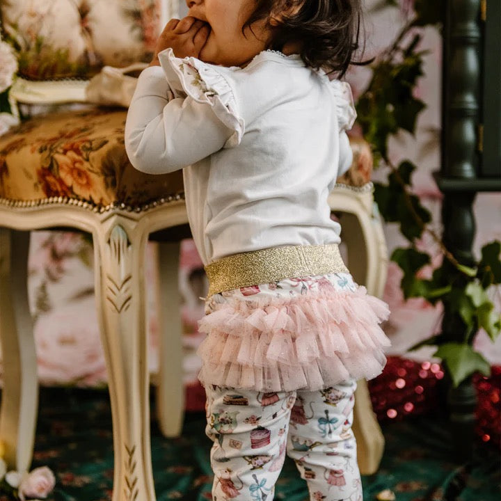Toddler wearing white top and patterned leggings with gold waistband and pink frill tutu detail at back