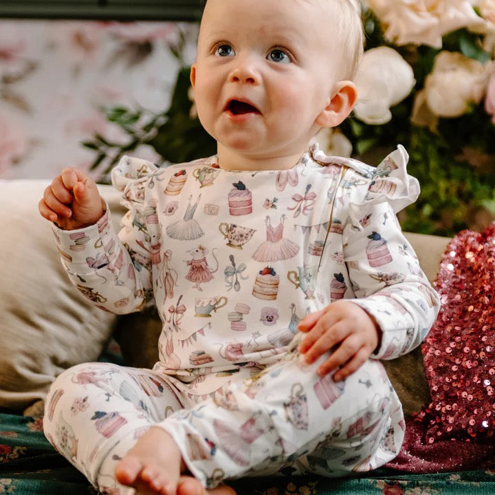 Baby sitting wearing frill coverall decorated with pastel illustrations of ballerina dresses cakes teacups and mice