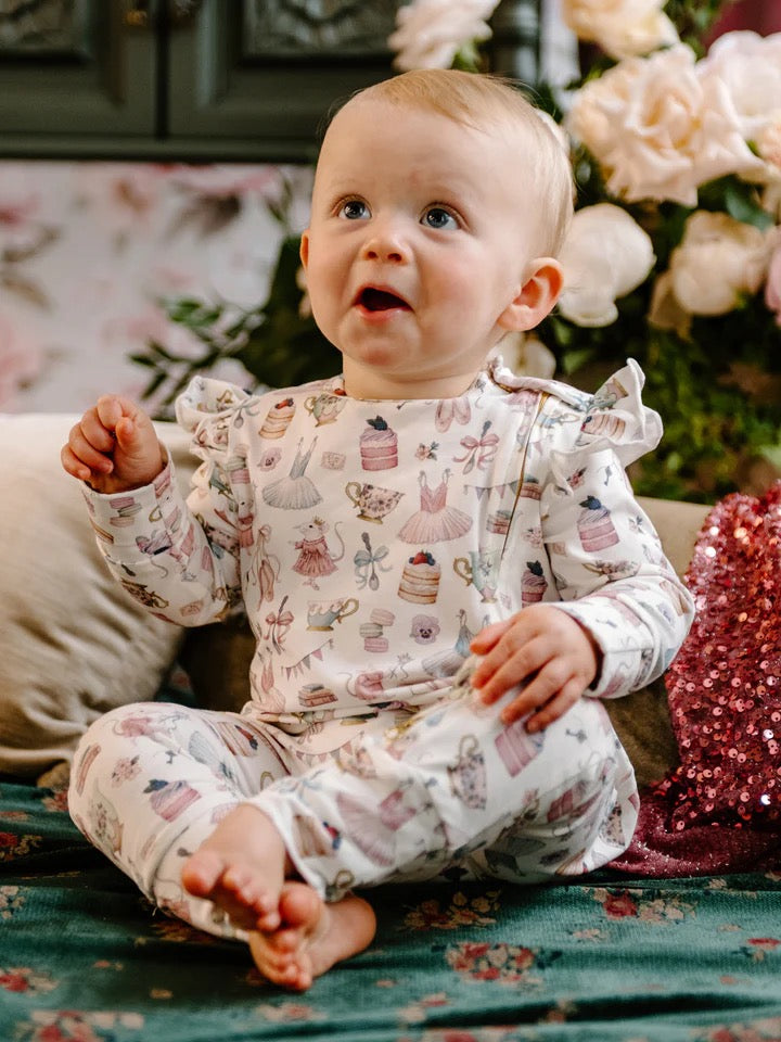 Baby sitting wearing frill coverall decorated with pastel illustrations of ballerina dresses cakes teacups and mice