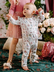 Baby standing wearing long sleeve frill coverall with whimsical party themed print including dresses cakes teacups and bows