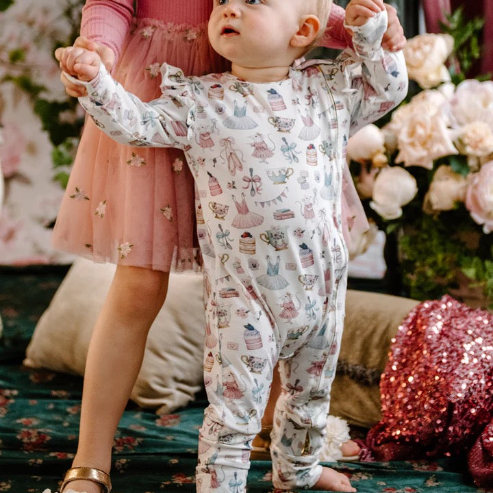 Baby standing wearing long sleeve frill coverall with whimsical party themed print including dresses cakes teacups and bows