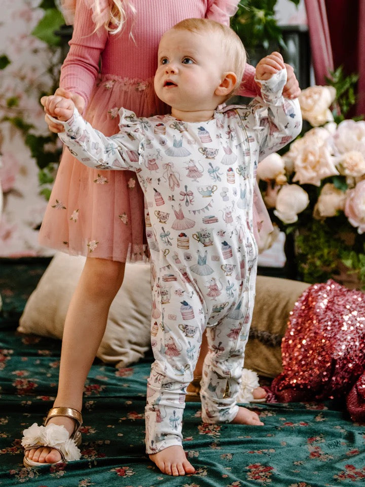 Baby standing wearing long sleeve frill coverall with whimsical party themed print including dresses cakes teacups and bows