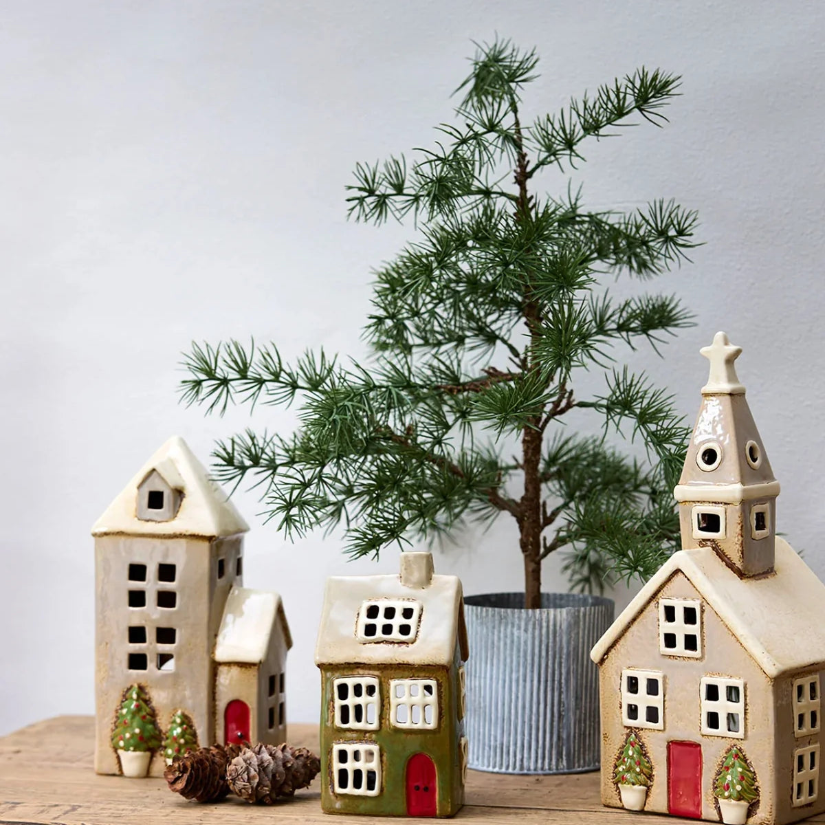 Christmas stone tea light houses including a beige ceramic church with red door and potted Christmas trees, styled with pine décor