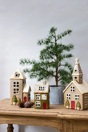 Christmas stone tea light houses including a beige ceramic church with red door and potted Christmas trees, styled with pine décor