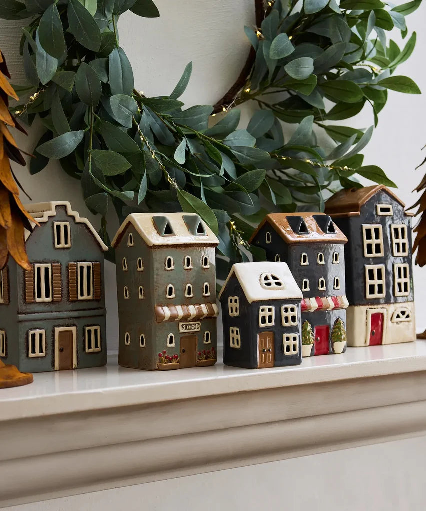 Collection of ceramic tea light houses styled on a mantel with greenery, featuring navy house with white roof in the center