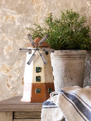 Decorative ceramic windmill tea light house displayed on a wooden table with rustic planter background