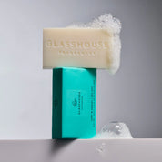 A turquoise GLASSHOUSE Fragrances "Lost In Amalfi / Sea Mist" 180g body bar with the cream-colored soap resting on top, covered in foamy soap bubbles.