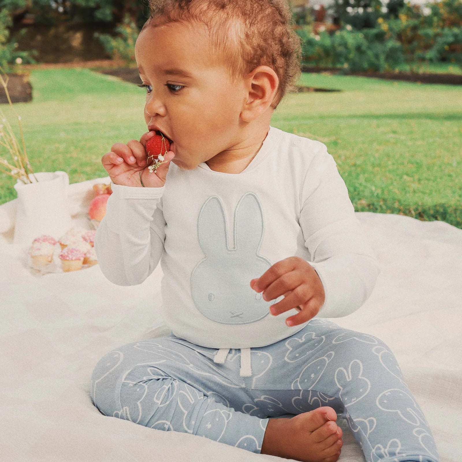 Baby sitting on picnic blanket wearing blue Miffy applique bodysuit and holding a strawberry