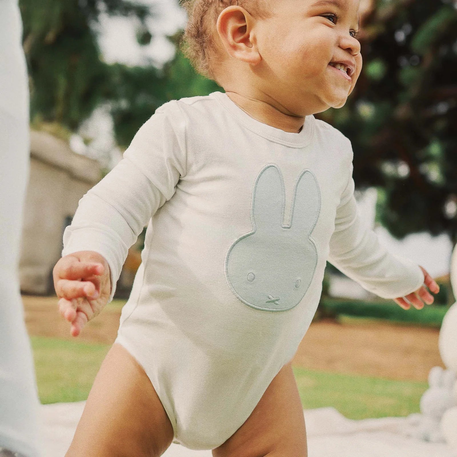 Baby wearing white Miffy bodysuit in blue applique while standing outside and smiling