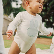 Baby wearing white Miffy bodysuit in blue applique while standing outside and smiling