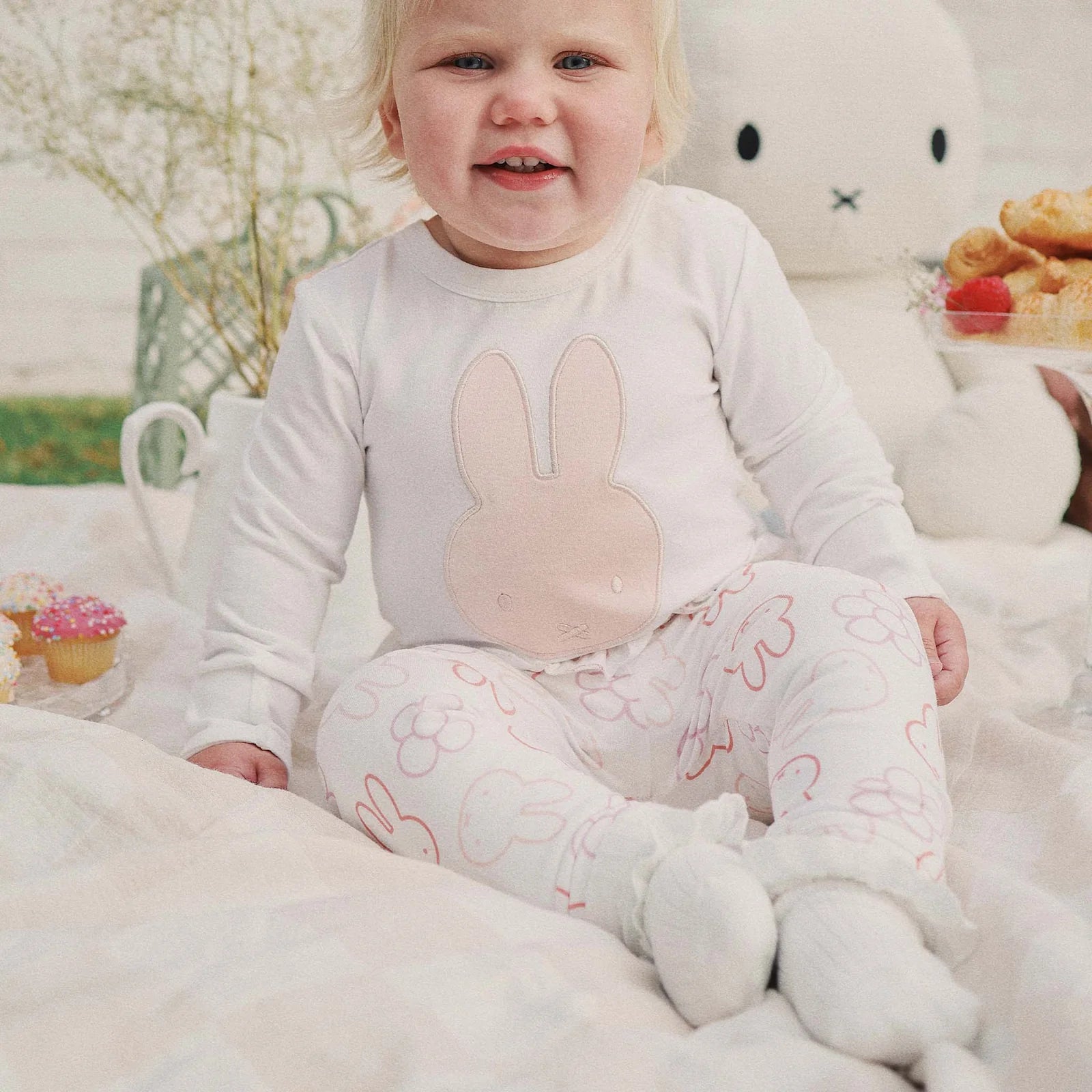 Baby sitting on a blanket in the Miffy bodysuit paired with pink printed leggings and snacks beside them.