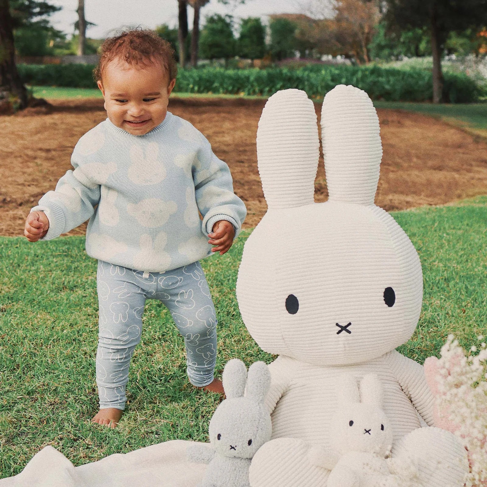 Toddler wearing Kynd Baby Miggy Leggings in moody blue with Miffy bunny print, standing on grass beside large Miffy plush.