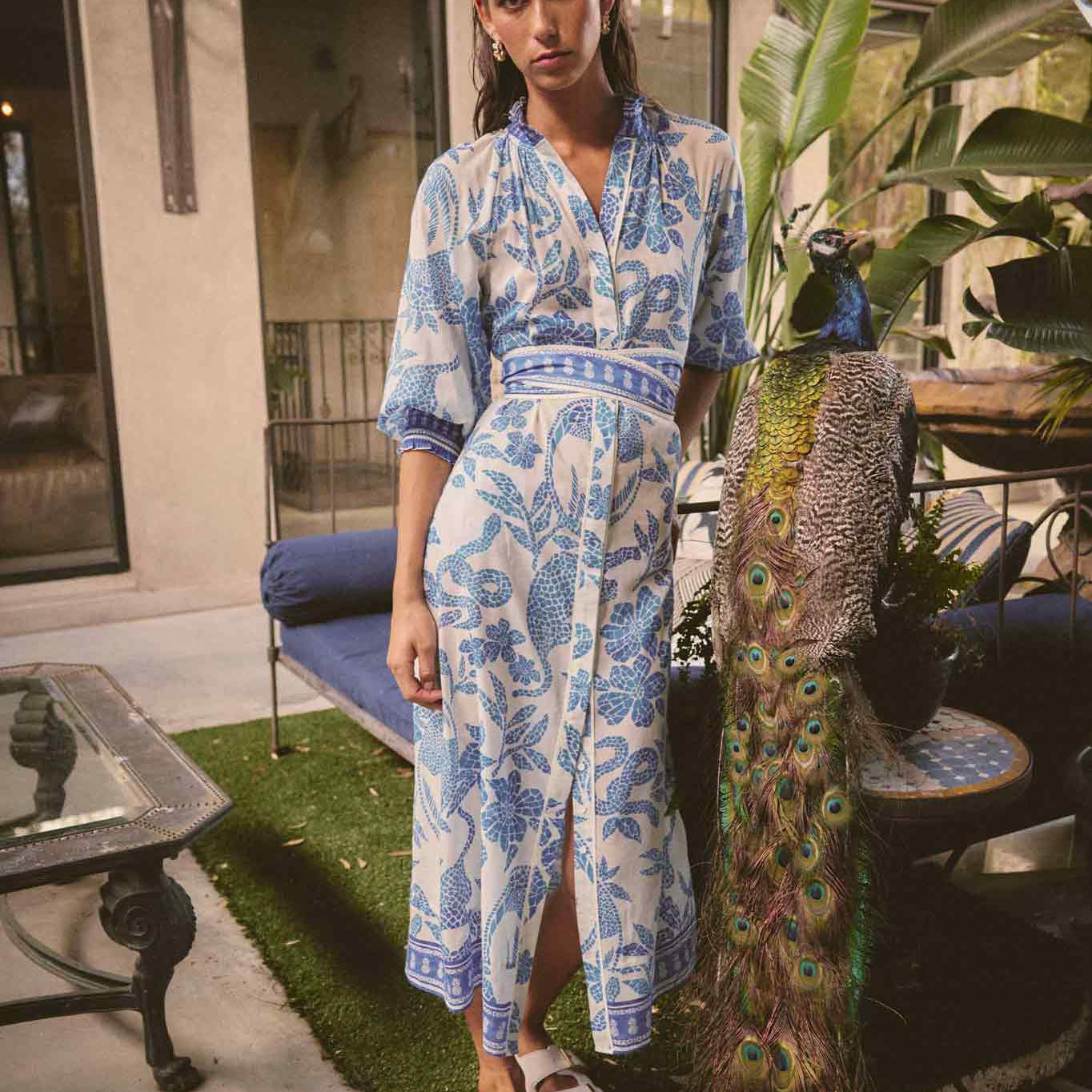 Model wearing ONCE WAS Carmen Silk Cotton Maxi Dress in Azure Mosaic with blue floral print, standing outdoors beside a peacock.