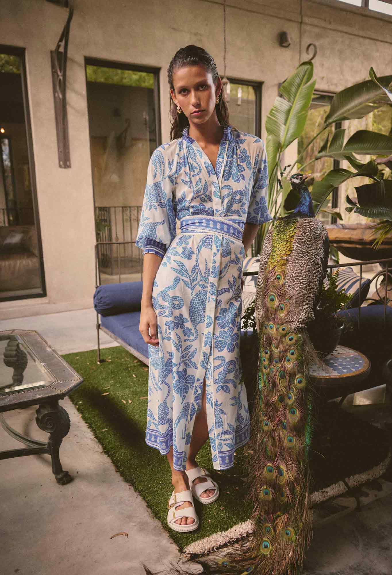 Model wearing ONCE WAS Carmen Silk Cotton Maxi Dress in Azure Mosaic with blue floral print, standing outdoors beside a peacock.