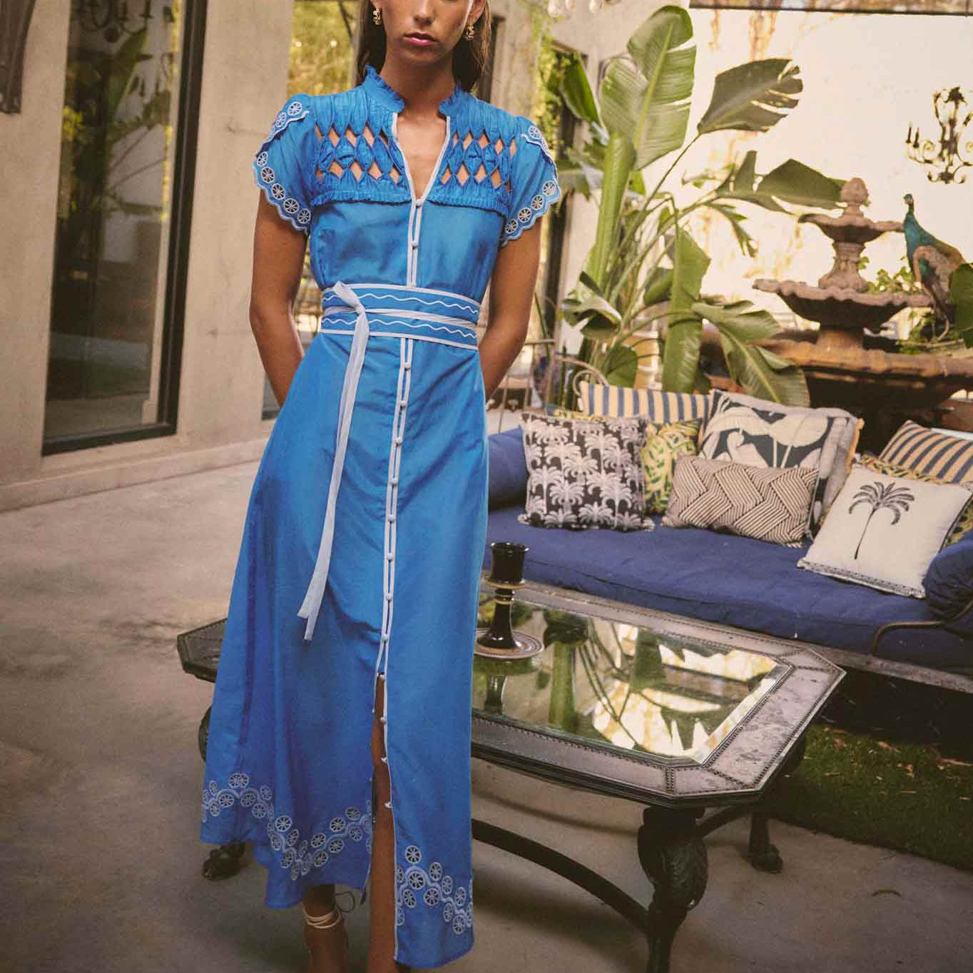 Model wearing ONCE WAS Xanthia Diamond Smock Yoke Silk Cotton Embroidered Dress in Blue Topaz, photographed outdoors with lounge and chandelier in background.
