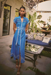 Model wearing ONCE WAS Xanthia Diamond Smock Yoke Silk Cotton Embroidered Dress in Blue Topaz, photographed outdoors with lounge and chandelier in background.