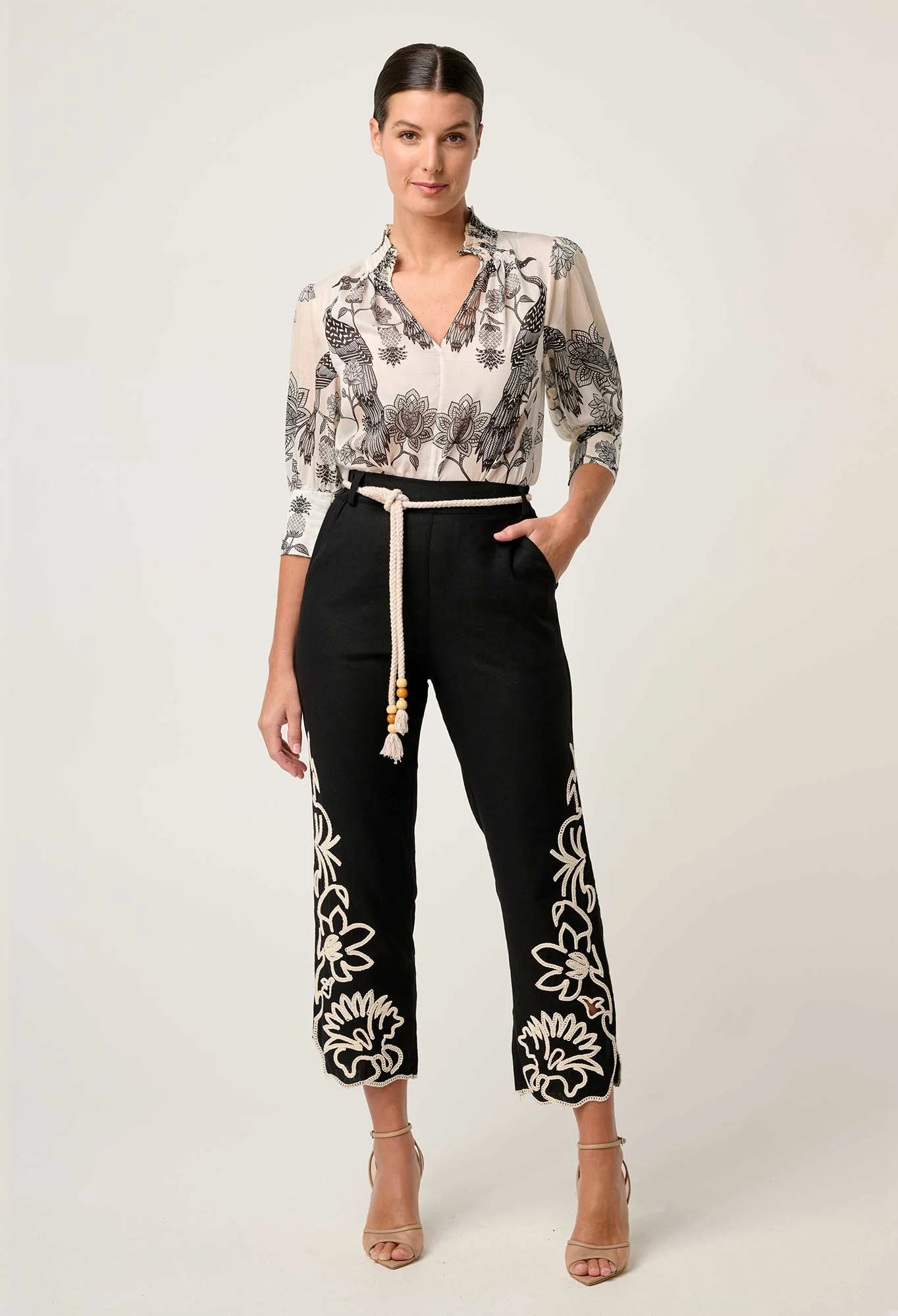 Front view of cream blouse with black floral bird print tucked into black pants with rope belt